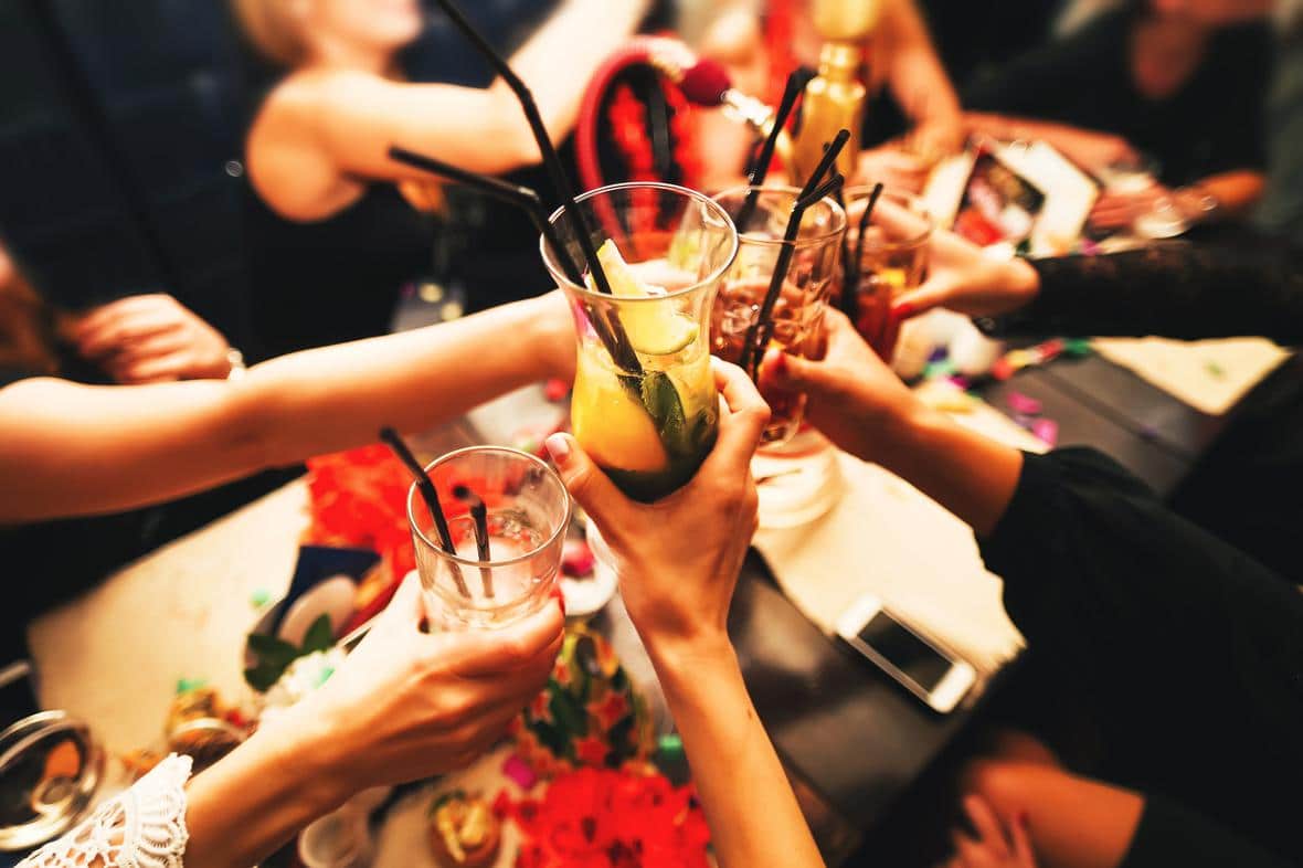 Group of friends cheering with colorful drinks on a wooden table in a dark setting