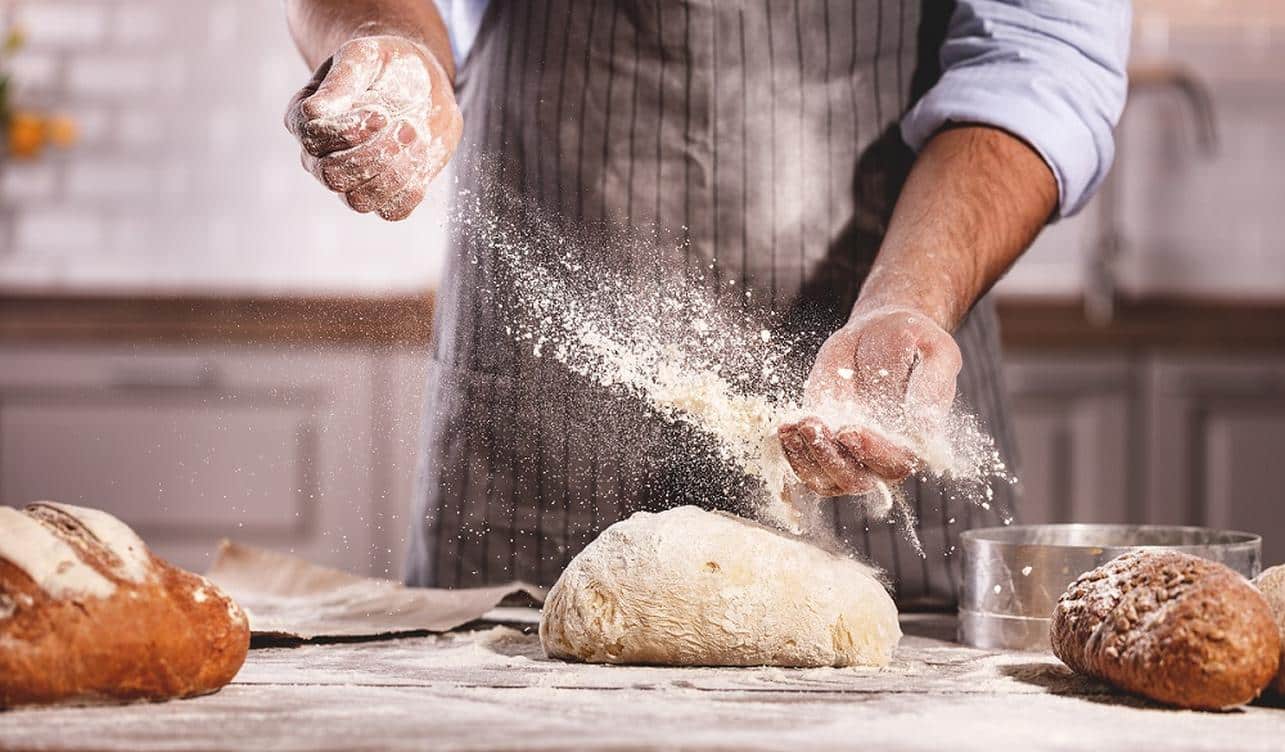 An expert baker is preparing bread and using flour on a grey table
