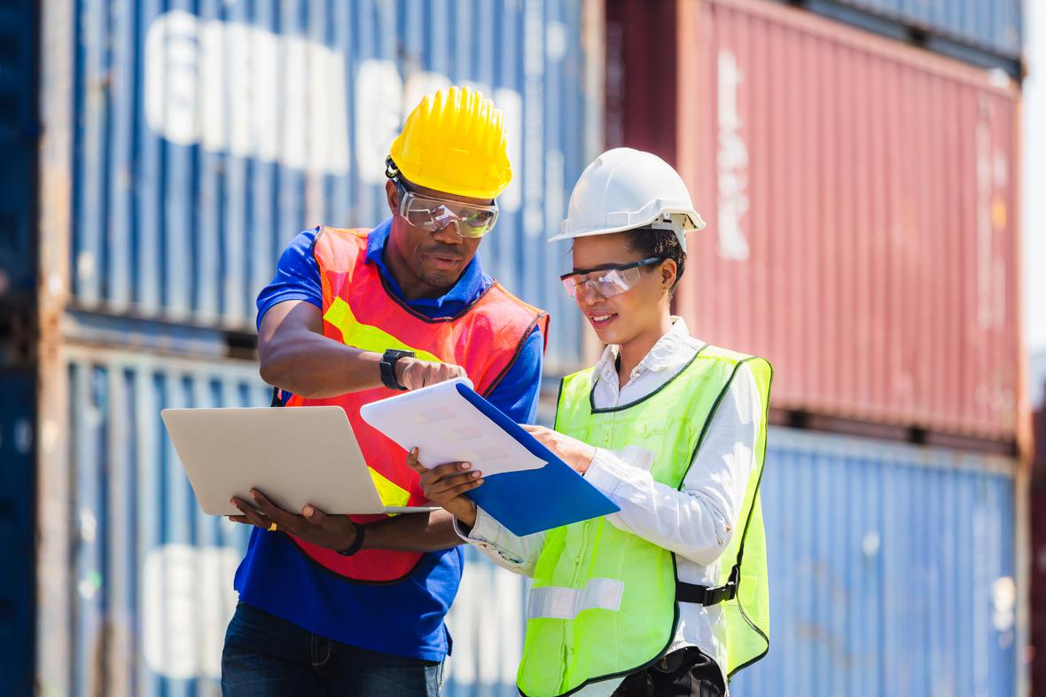 Female foreman safety vest using clipboard checklist and Worker man in hardhat holding laptop for control loading containers box from cargo AdobeStock 329816941