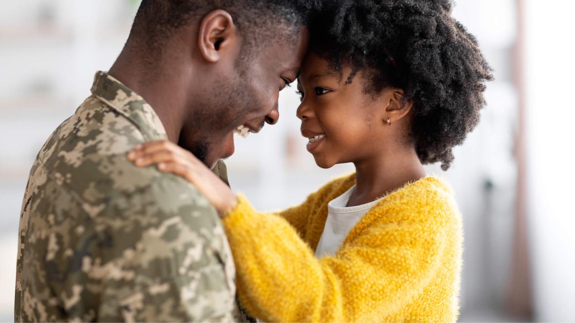 A person in military fatigues smiling and putting their forehead against the forehead of a smiling child
