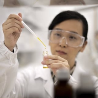A person wearing lab goggles uses a dropper to add liquid to a small container