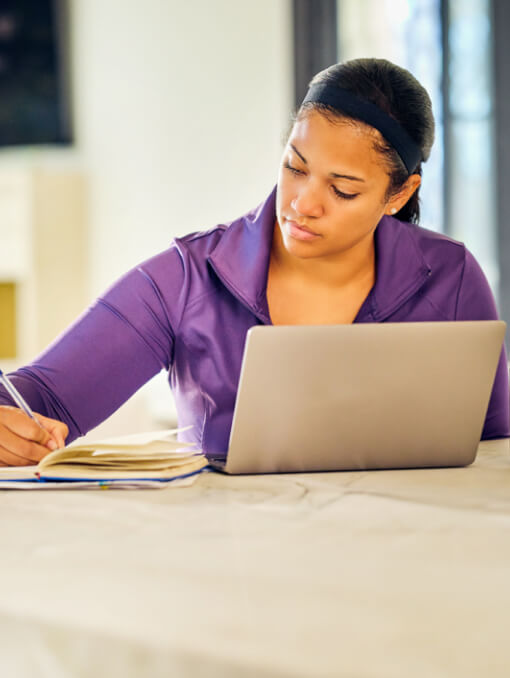A person sits at table while writing in a notebook next to a laptop