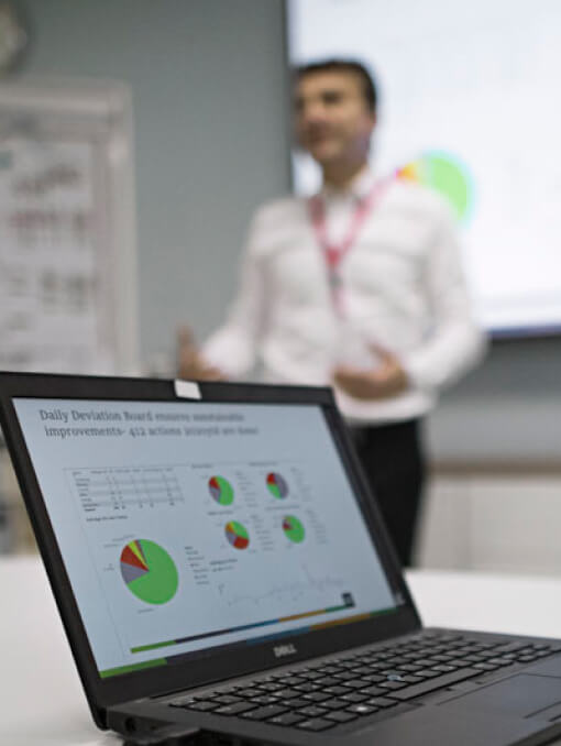 Closeup of a laptop on a table with a person presenting in front of a screen in the background