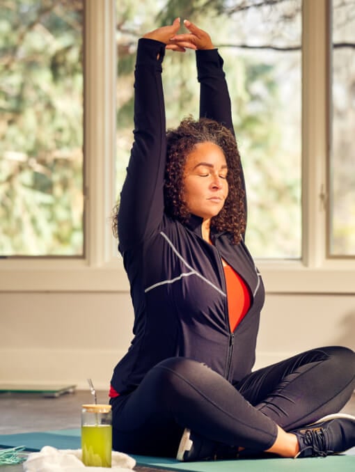 A person sits on a yoga mat with their legs crossed and arms stretched upward with a sports drink on the floor next to them