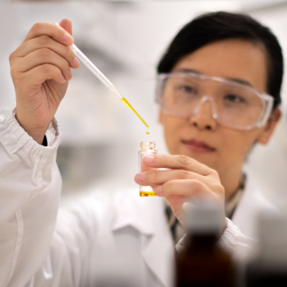 A close-up of a female scientist wearing a lab coat and goggles, using a dropper to add liquid to a test tube.