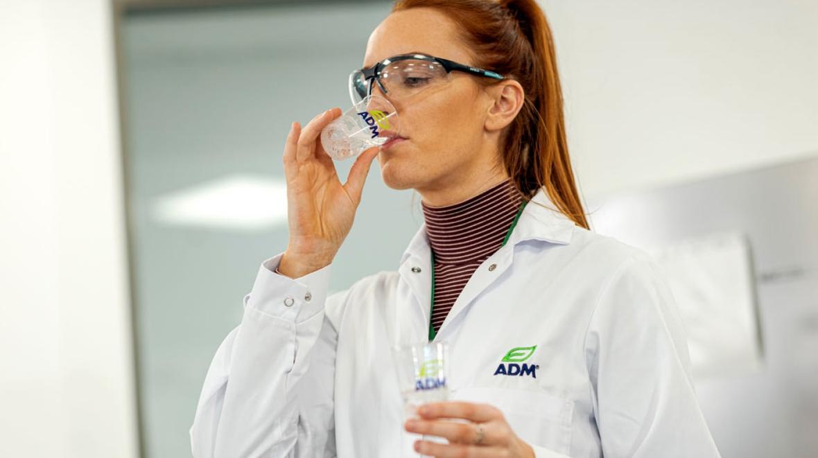 An ADM employee in a lab coat and safety glasses drinks from a small ADM-branded cup