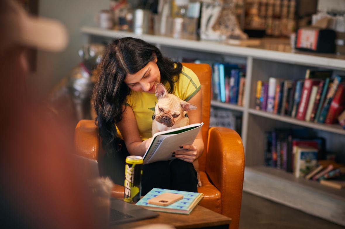 Woman sitting on orange chair and holding small dog and notebook with bookshelf behind Palm Springs 05942 RT V01 231112 lores