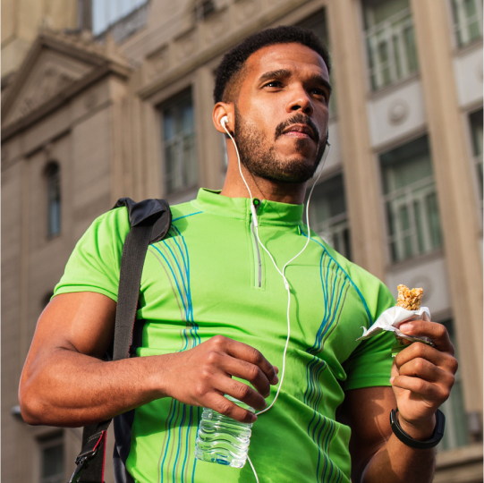 A man walks down the street looking like he just left the gym and is now enjoying a bottle of water and reduced sugar nutritional bar.