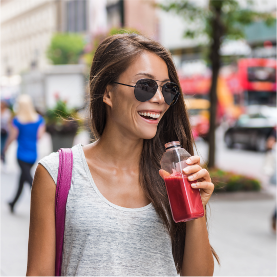  A woman walking down a city street in sunglasses is smiling as she enjoys a reduced sugar fruit smoothie in a bottle.