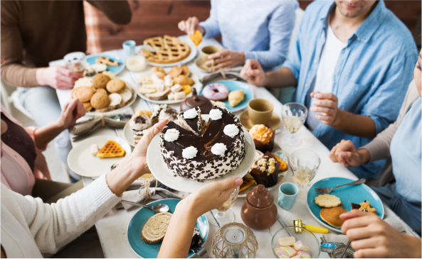  A group of friends sit around a table piled high with sugar reduced desserts passing plates amongst each other.