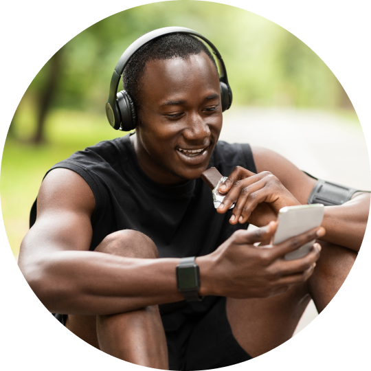 A man sitting on the ground wearing headphones and looking at his phone while eating a protein bar.