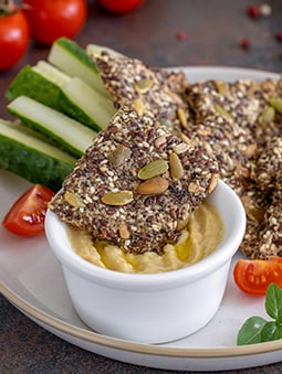 A plate of food with a bowl of hummus, seeded crackers, and vegetables