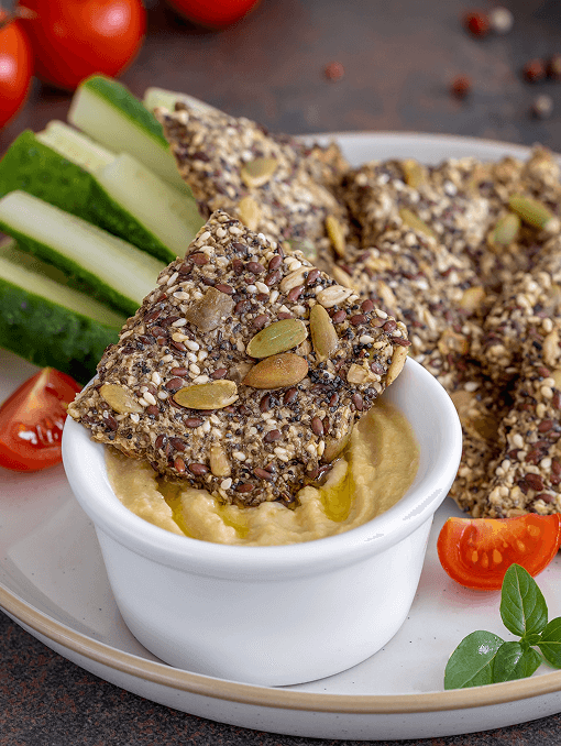 A plate of food with a bowl of hummus, seeded crackers, and vegetables