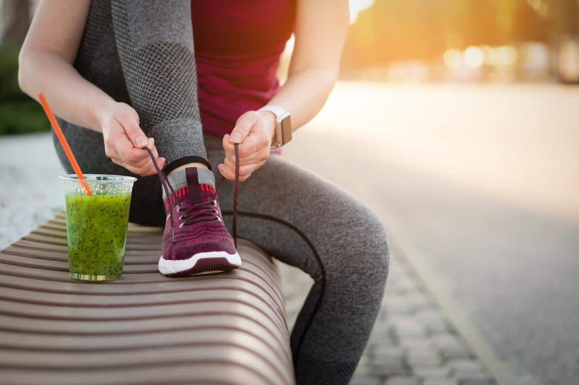 Green detox smoothie cup and woman lacing running shoes on public bench