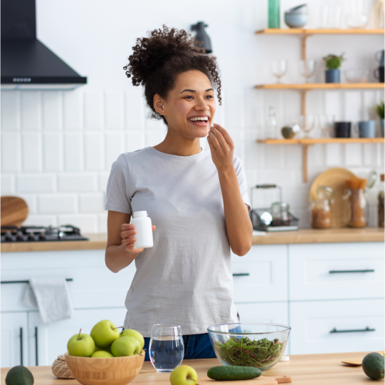 A woman eating a plant based botanical extract from a white bottle in the kitchen with a bowl of green apples, leafy greens and a glass of water in front of her.
