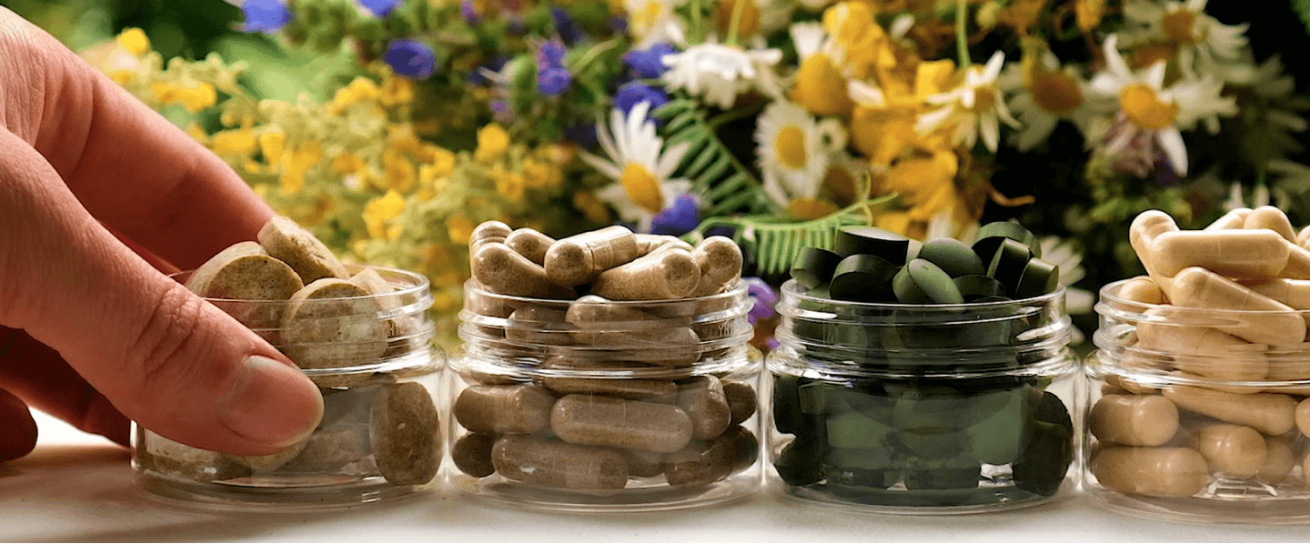 A close-up of botanical extracts plant based pills kept in small glass jars on a table with colourful plants in the background.