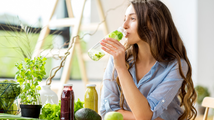 A woman drinking fruits infused water at the table surrounded by plants, plant based juices and fruits.