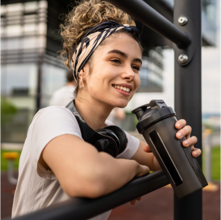 A woman with a protein drink in her hand and an over-the-ear headphone around her neck leaning against a railing.