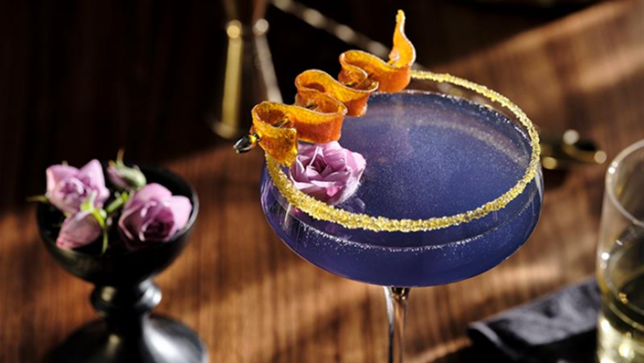 A blue-purple cocktail in a coupe glass with garnishes