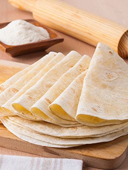 A stack of folded flour tortillas next to a rolling pin and a bowl of flour