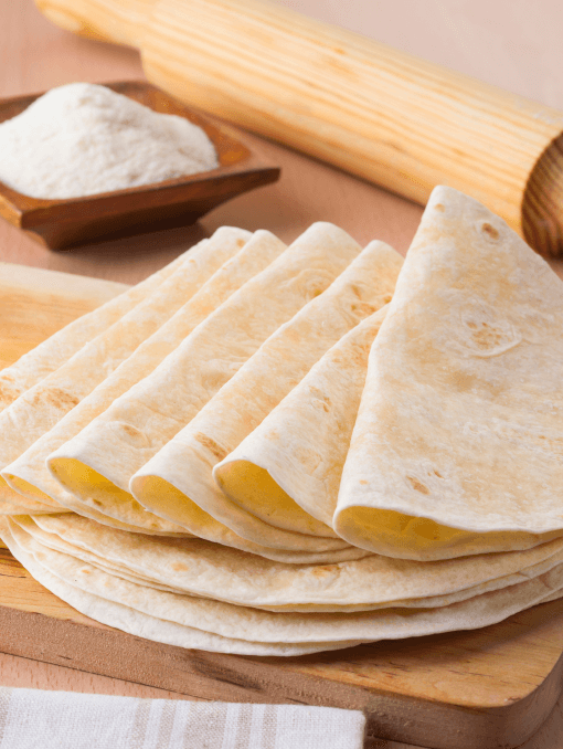 A stack of folded flour tortillas next to a rolling pin and a bowl of flour