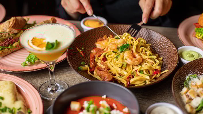 A person sitting at a table full of drinks and plates, enjoying a pasta dish.