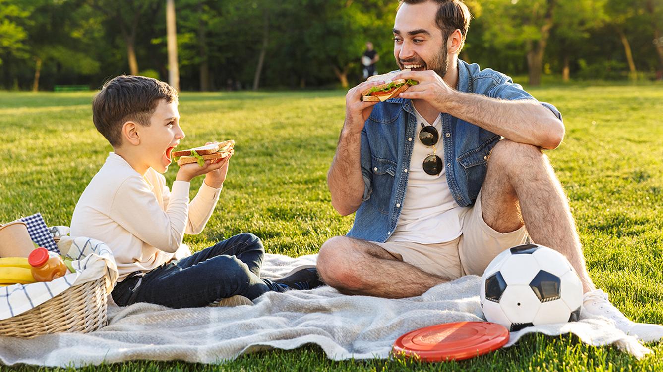 A person and a child eating sandwiches on a picnic blanket