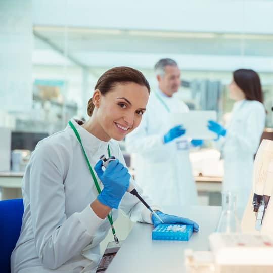 A woman scientist with a white coat performs an experiment in a lab with 2 scientists in the background