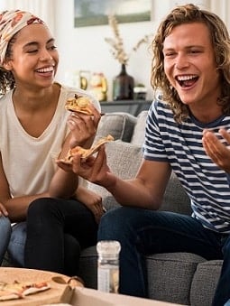 A couple eats pizza on a sofa while smiling and laughing