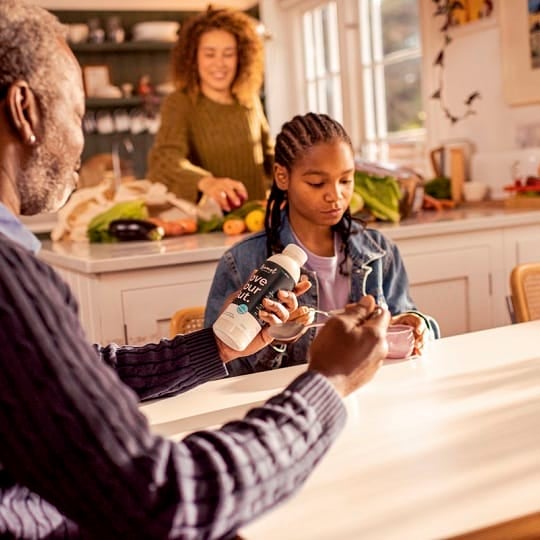 A young black girl has breakfast while her dad looks at a bottle of dietary supplements