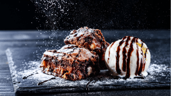 A fancy black plate sits on a black wood table holding two chocolate brownies and a scoop of vanilla ice cream drizzled in chocolate sauce while someone out of frame sprinkles powder sugar on top.