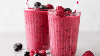 Two berry smoothies in glasses with straws have fresh berries sprinkled on top of them, and a few more berries have fallen onto the white table beside them.