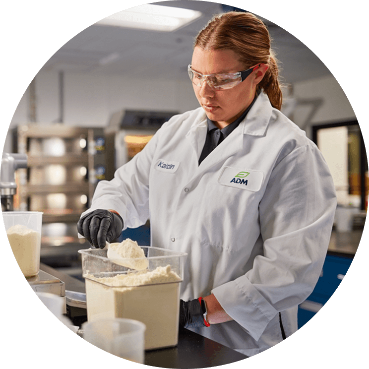 An ADM employee in a lab coat and safety goggles scooping powder out of a container