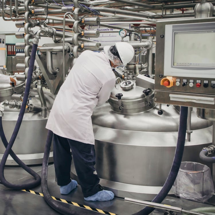 An employee in a white lab coat and hardhat in an industrial scientific setting
