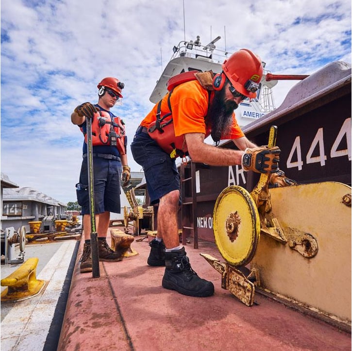 Two workers in orange safety gear, life vests, and red hard hats operate machinery on the deck of a barge, with one worker turning a large yellow wheel mechanism as another looks on holding a tool.