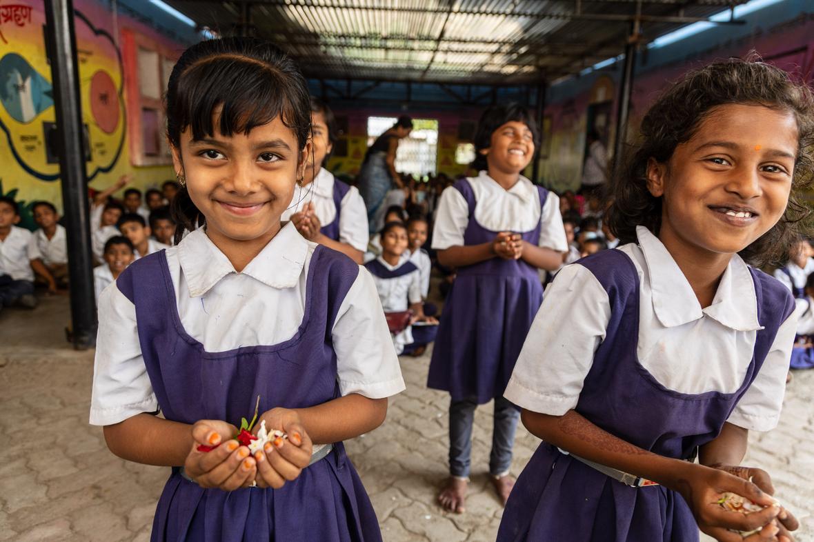 A young girl in a white and purple school uniform smiles at the camera inside a colorfully painted classroom, with other uniformed students visible in the background.