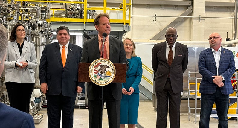 A group of people wearing suits in a factory setting. A man is speaking behind a lectern.