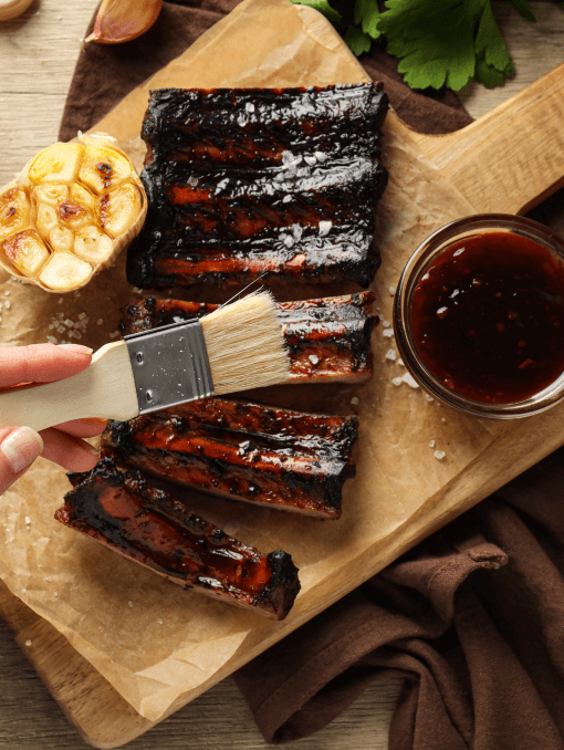A person’s hand is shown brushing a dark brown marinade onto ribs