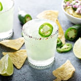 Margaritas made with agave sit on a table with tortilla chips and jalapeno slices 