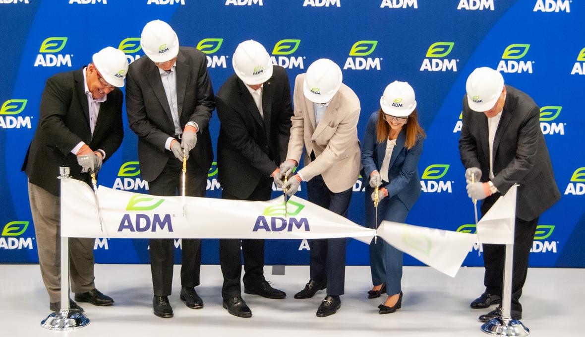 A group of people wearing suits and hard hats in front of a blue backdrop with the ADM logo. They are using large scissors to cut a ceremonial ribbon banner with the ADM logo.
