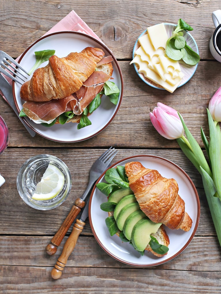 Two white plates with croissant sandwiches, with slices of cheese and lemons on the side.