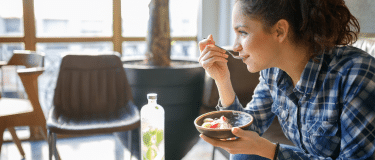 A woman sitting on a couch eating breakfast from a bowl in her hand while her coffee and bottle of water sit on the table in front of her.