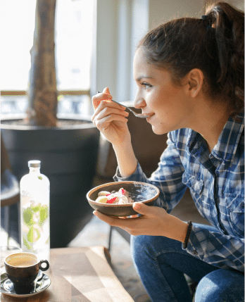 A woman sitting on a couch eating breakfast from a bowl in her hand while her coffee and bottle of water sit on the table in front of her.