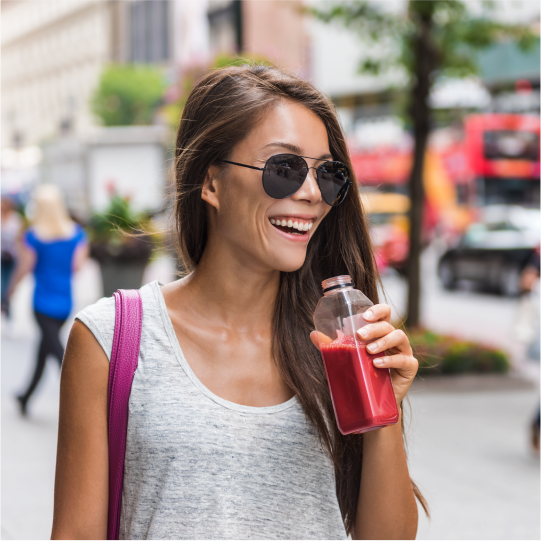 A woman walking down a city street in sunglasses is smiling as she enjoys a reduced sugar fruit smoothie in a bottle.