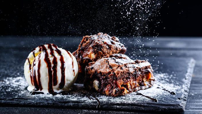 A close up of a stylized plate of reduced sugar brownies and ice cream on a plate being sprinkled with powdered sugar.