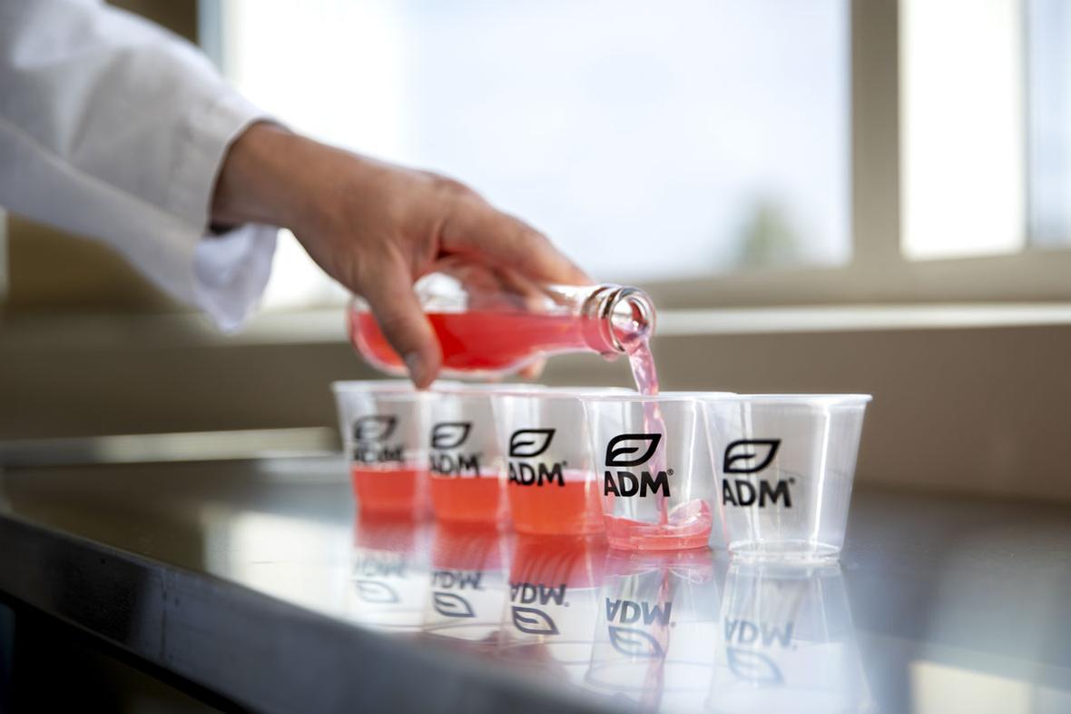 A pink reduced sugar soda being poured into a line of several small ADM branded tasting glasses.