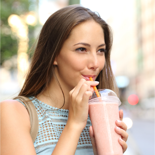A woman stands outside on a city street sipping a reduced sugar smoothie through a straw.