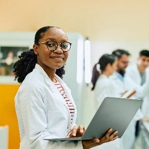 A black scientist in a white lab coat smiles while on her PC