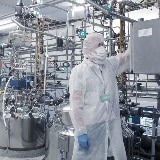 A man in a white lab coat is in a factory with many iron tubes