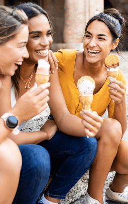 Three young women smiling while sitting on a curb enjoying sugar-free ice cream cones.